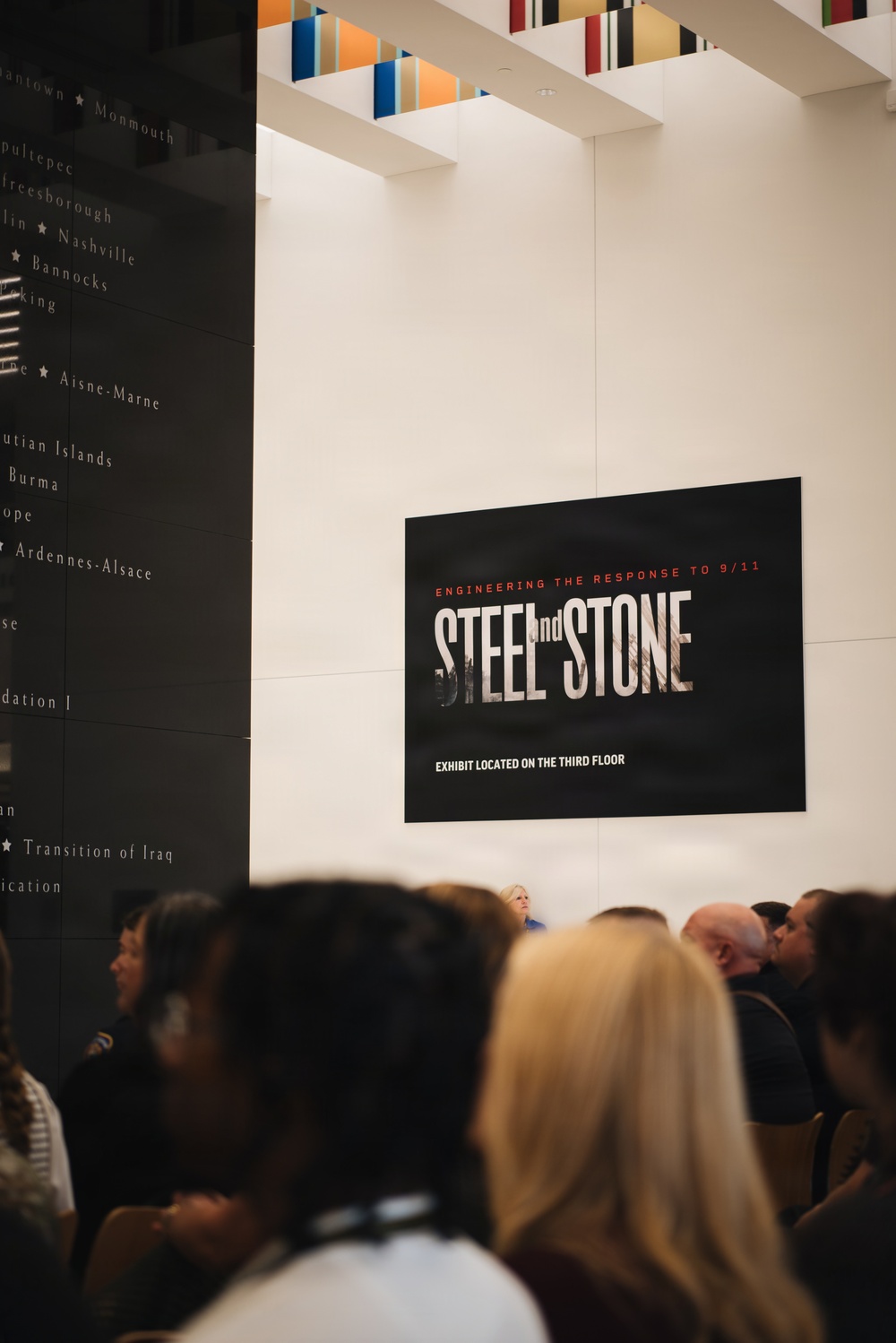 The image shows a museum or memorial setting with a prominent exhibit sign reading 'Steel and Stone: Engineering the Response to 9/11,' with a note that the exhibit is located on the third floor. A crowd of visitors is gathered in the foreground, slightly out of focus. To the left is a dark panel listing historical battle names and military campaigns.