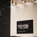 The image shows a museum or memorial setting with a prominent exhibit sign reading 'Steel and Stone: Engineering the Response to 9/11,' with a note that the exhibit is located on the third floor. A crowd of visitors is gathered in the foreground, slightly out of focus. To the left is a dark panel listing historical battle names and military campaigns.