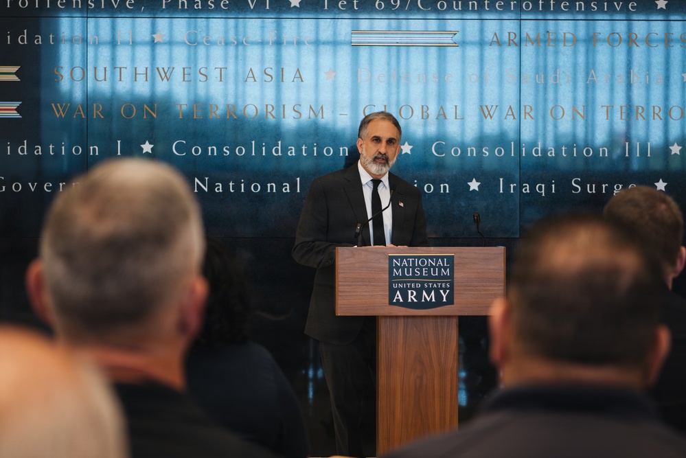 A man in a dark suit with a gray beard is speaking at a podium inside the National Museum of the United States Army. The background wall displays text referencing various military campaigns and conflicts including 'War on Terrorism,' 'Southwest Asia,' and 'Iraqi Surge.' An audience is visible in the foreground, seated with their backs to the camera.