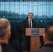 A man in a dark suit with a gray beard is speaking at a podium inside the National Museum of the United States Army. The background wall displays text referencing various military campaigns and conflicts including 'War on Terrorism,' 'Southwest Asia,' and 'Iraqi Surge.' An audience is visible in the foreground, seated with their backs to the camera.