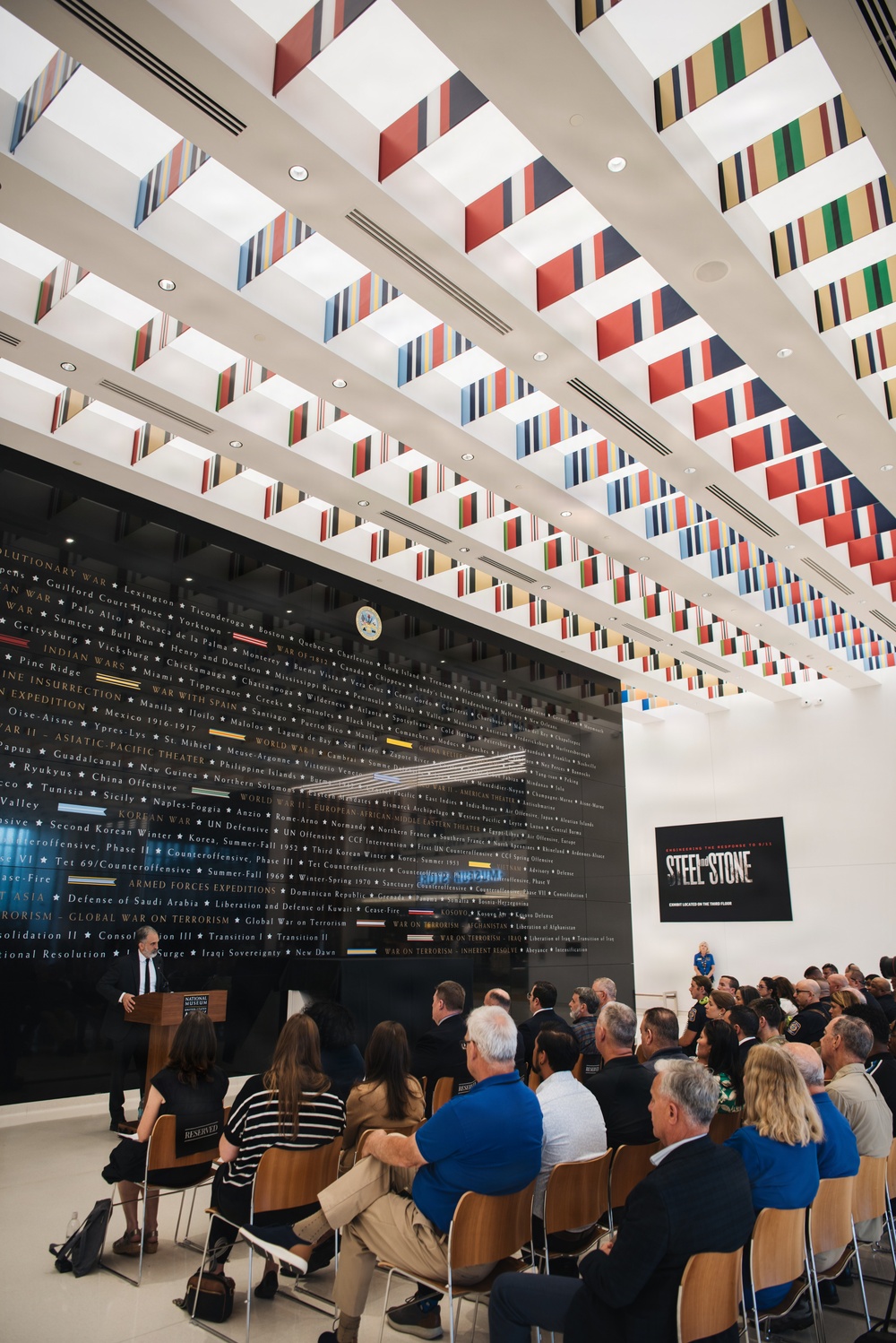 A formal presentation or ceremony is taking place inside what appears to be the National Museum of the United States Army, featuring a large black wall displaying U.S. Army campaign and battle names across American military history. A speaker stands at a podium on the left while an audience of approximately 50-75 people is seated facing him. A sign on the right wall advertises an exhibit titled 'Steel on Stone,' described as being located on the third floor.