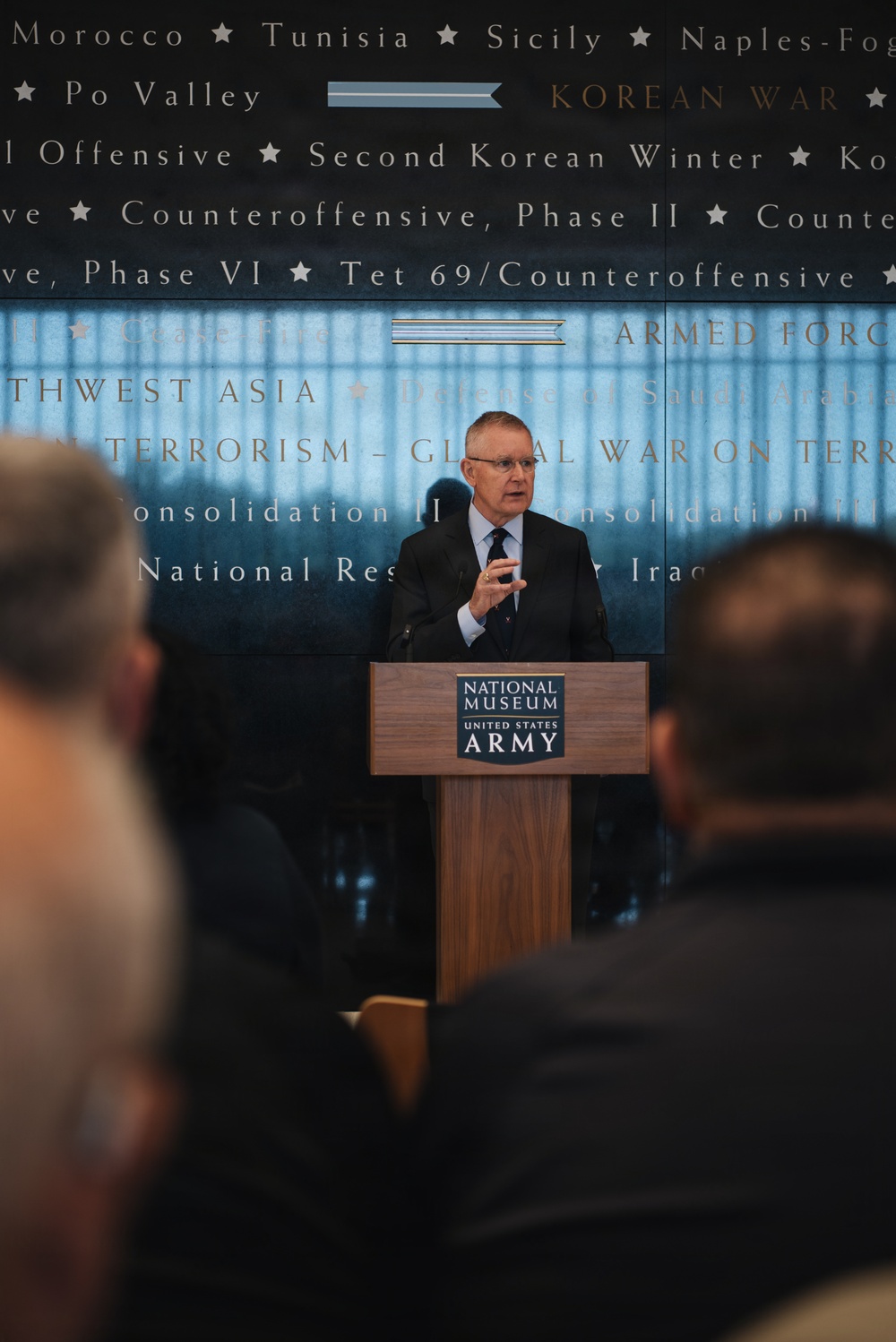 A man in a dark suit is speaking at a podium bearing the 'National Museum United States Army' logo. He is addressing an audience, with several attendees visible in the foreground as silhouettes. The background wall displays a chronological list of U.S. military campaigns and conflicts, ranging from World War II battles to the Global War on Terrorism.