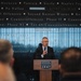 A man in a dark suit is speaking at a podium bearing the 'National Museum United States Army' logo. He is addressing an audience, with several attendees visible in the foreground as silhouettes. The background wall displays a chronological list of U.S. military campaigns and conflicts, ranging from World War II battles to the Global War on Terrorism.