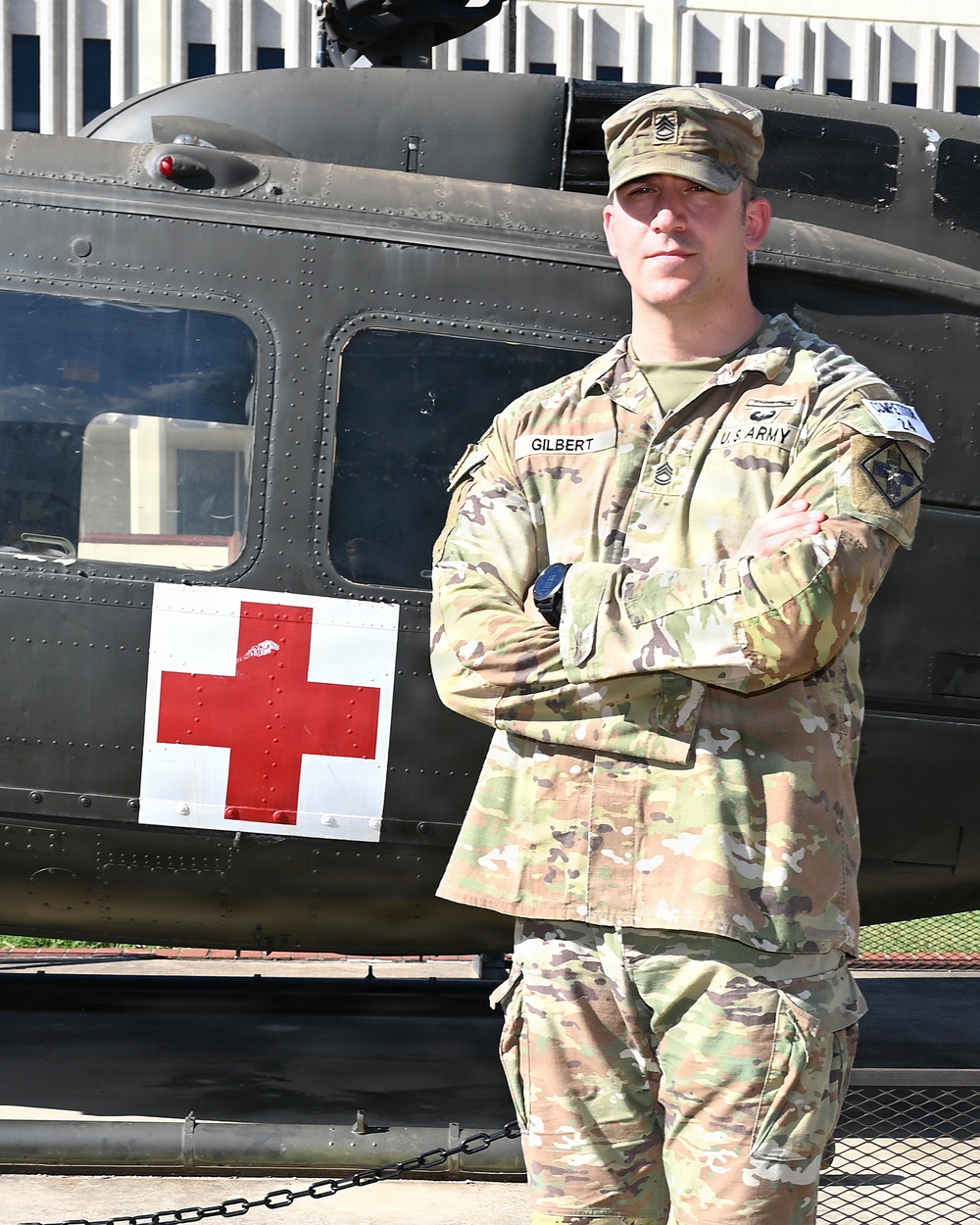 A U.S. Army soldier in OCP (Operational Camouflage Pattern) uniform stands with arms crossed in front of a dark military medical helicopter marked with a red cross. The soldier's name tape reads 'GILBERT' and the branch tape reads 'U.S. ARMY,' with rank insignia visible on the cap consistent with a Staff Sergeant or Sergeant First Class. The image appears to be an official or semi-official portrait-style photograph taken outdoors.