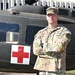 A U.S. Army soldier in OCP (Operational Camouflage Pattern) uniform stands with arms crossed in front of a dark military medical helicopter marked with a red cross. The soldier's name tape reads 'GILBERT' and the branch tape reads 'U.S. ARMY,' with rank insignia visible on the cap consistent with a Staff Sergeant or Sergeant First Class. The image appears to be an official or semi-official portrait-style photograph taken outdoors.