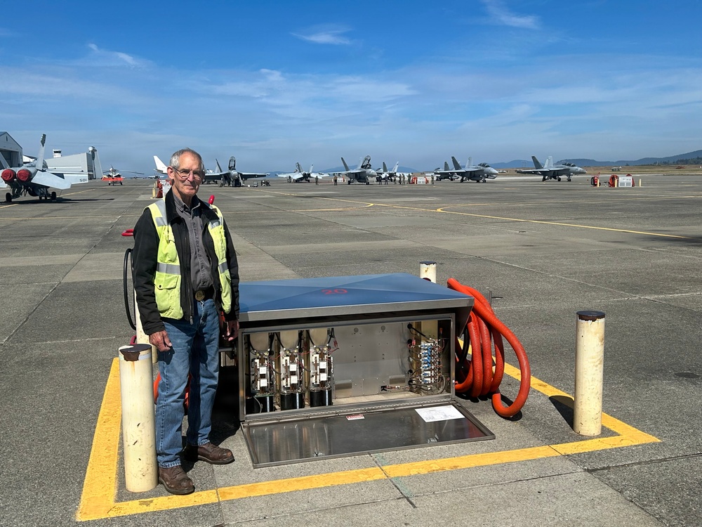 A man wearing a yellow high-visibility safety vest stands on a military airfield tarmac next to an open ground service equipment panel or utility pit, which contains electrical or pneumatic components and an orange flexible hose. Multiple military jet aircraft, appearing to be F/A-18 Hornets or Super Hornets, are visible lined up on the flight line in the background. The setting appears to be a naval air station or military air base.