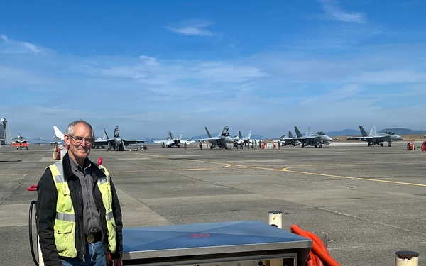 A man wearing a yellow high-visibility safety vest stands on a military airfield tarmac next to an open ground service equipment panel or utility pit, which contains electrical or pneumatic components and an orange flexible hose. Multiple military jet aircraft, appearing to be F/A-18 Hornets or Super Hornets, are visible lined up on the flight line in the background. The setting appears to be a naval air station or military air base.