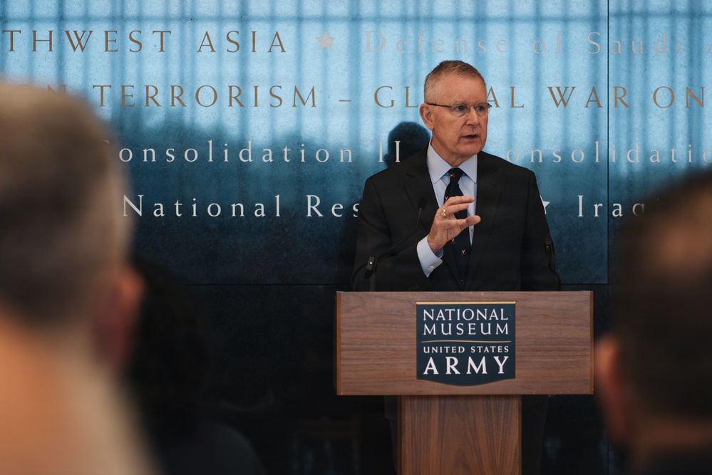 A man in a dark suit and tie is speaking at a podium bearing the 'National Museum of the United States Army' insignia. He appears to be delivering a formal address or presentation to an audience, with a large illuminated backdrop displaying text referencing military campaigns including 'Global War on Terrorism,' 'Southwest Asia,' and 'Defense of Saudi.' The setting is an official ceremonial or commemorative event at the museum.