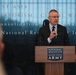 A man in a dark suit and tie is speaking at a podium bearing the 'National Museum of the United States Army' insignia. He appears to be delivering a formal address or presentation to an audience, with a large illuminated backdrop displaying text referencing military campaigns including 'Global War on Terrorism,' 'Southwest Asia,' and 'Defense of Saudi.' The setting is an official ceremonial or commemorative event at the museum.