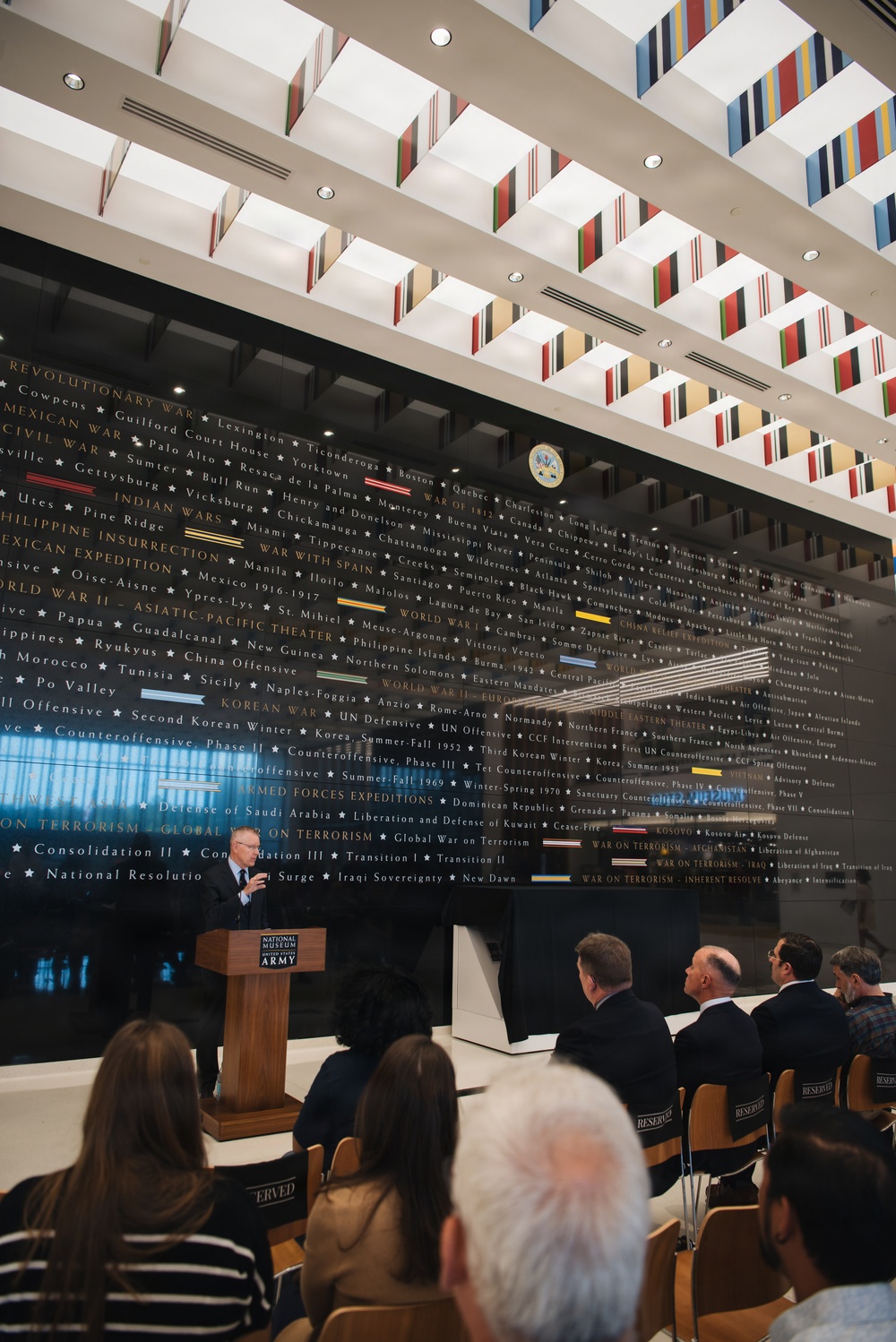A formal speaking event is taking place at the National Museum of the United States Army, with a speaker at a wooden podium addressing a seated audience. Behind the speaker is a large dark wall displaying the chronological history of U.S. Army campaigns and battles, from the Revolutionary War through modern conflicts. The ceiling features a distinctive architectural design with colorful military campaign ribbon-inspired decorative elements.