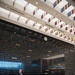 A formal speaking event is taking place at the National Museum of the United States Army, with a speaker at a wooden podium addressing a seated audience. Behind the speaker is a large dark wall displaying the chronological history of U.S. Army campaigns and battles, from the Revolutionary War through modern conflicts. The ceiling features a distinctive architectural design with colorful military campaign ribbon-inspired decorative elements.