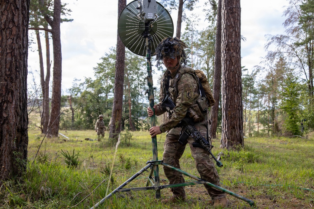 A soldier in full combat gear and camouflage uniform is setting up or adjusting a portable satellite/communications dish antenna mounted on a tripod in a forested clearing. The soldier is wearing a helmet with camouflage netting and carrying a rifle, with a second soldier visible in the background. The scene appears to be a military field exercise or training operation.