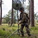 A soldier in full combat gear and camouflage uniform is setting up or adjusting a portable satellite/communications dish antenna mounted on a tripod in a forested clearing. The soldier is wearing a helmet with camouflage netting and carrying a rifle, with a second soldier visible in the background. The scene appears to be a military field exercise or training operation.
