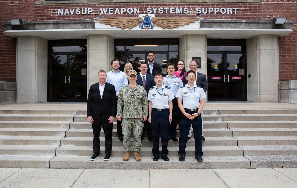 A group of approximately 12 individuals poses for a formal group photograph on the steps of the NAVSUP Weapon Systems Support building. The group includes personnel in U.S. Navy camouflage uniforms, what appear to be Japanese Maritime Self-Defense Force or similar Asian military uniforms in light blue/white, and civilian business attire. This appears to be an official visit or exchange program between military and civilian personnel.