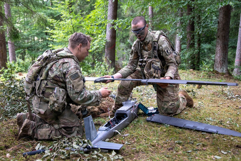 Two military personnel in MultiCam/OCP camouflage uniforms are assembling or preparing a fixed-wing unmanned aerial vehicle (UAV/drone) in a forested area. Both soldiers are kneeling on the ground and appear to be attaching wing components to the drone's fuselage. The setting suggests a field training exercise or operational deployment.