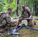 Two military personnel in MultiCam/OCP camouflage uniforms are assembling or preparing a fixed-wing unmanned aerial vehicle (UAV/drone) in a forested area. Both soldiers are kneeling on the ground and appear to be attaching wing components to the drone's fuselage. The setting suggests a field training exercise or operational deployment.