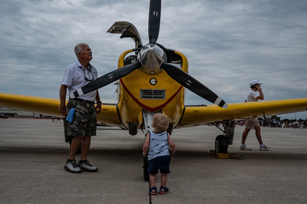 An outdoor airshow scene on a tarmac featuring a bright yellow vintage propeller aircraft as the centerpiece. An elderly man in a white polo shirt and camouflage shorts stands beside the aircraft's nose, while a toddler in a shark-print shirt looks up at the propeller. Additional visitors are visible in the background under an overcast sky.