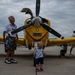 An outdoor airshow scene on a tarmac featuring a bright yellow vintage propeller aircraft as the centerpiece. An elderly man in a white polo shirt and camouflage shorts stands beside the aircraft's nose, while a toddler in a shark-print shirt looks up at the propeller. Additional visitors are visible in the background under an overcast sky.