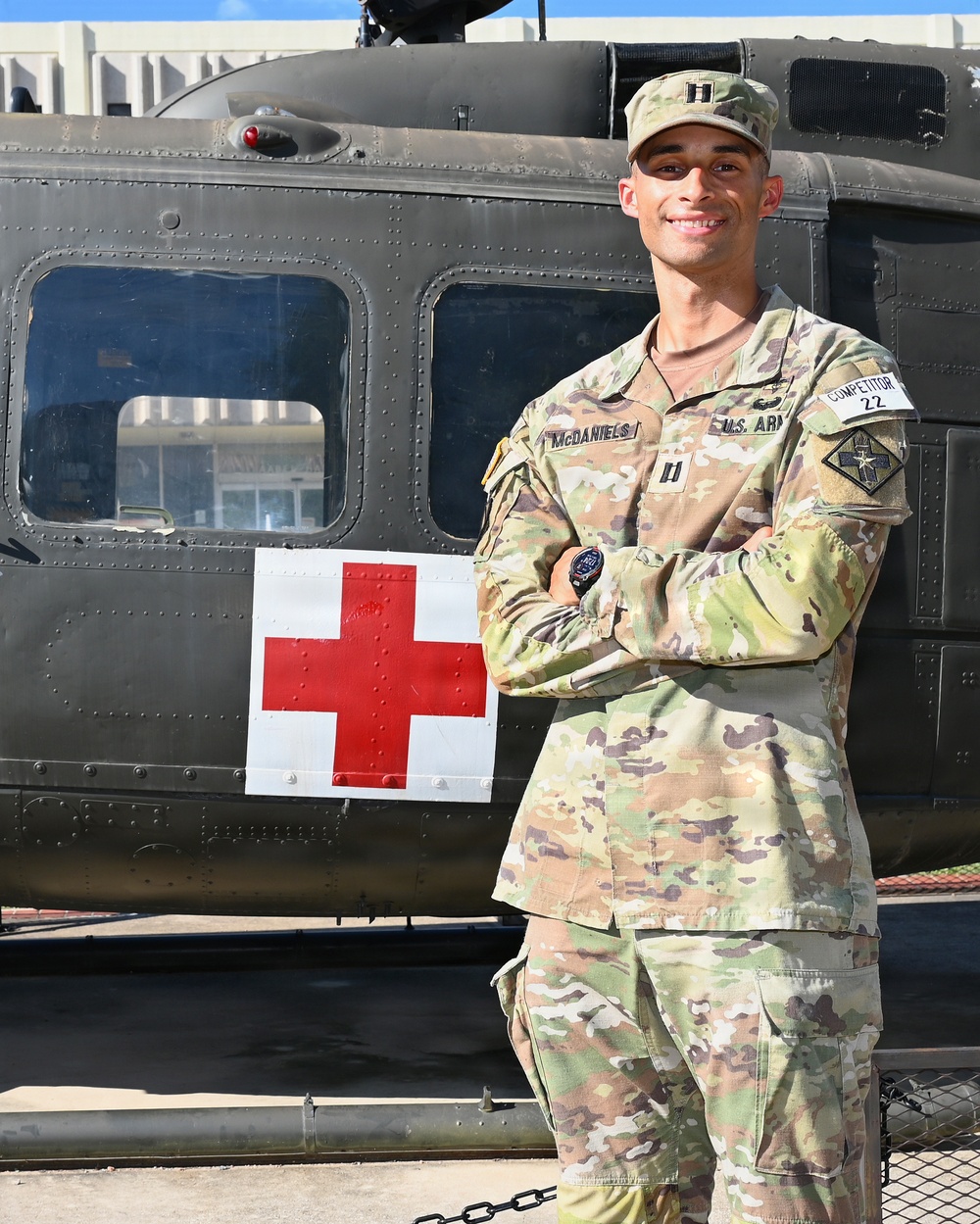 A U.S. Army Captain named McDaniels stands in front of a military medical (MEDEVAC) helicopter, identifiable by the prominent Red Cross marking on its fuselage. He is wearing OCP (Operational Camouflage Pattern) uniform with Captain rank insignia and a 'Competitor 22' armband, suggesting participation in a military competition. The photo appears to be an official or promotional portrait taken outdoors in clear daylight.