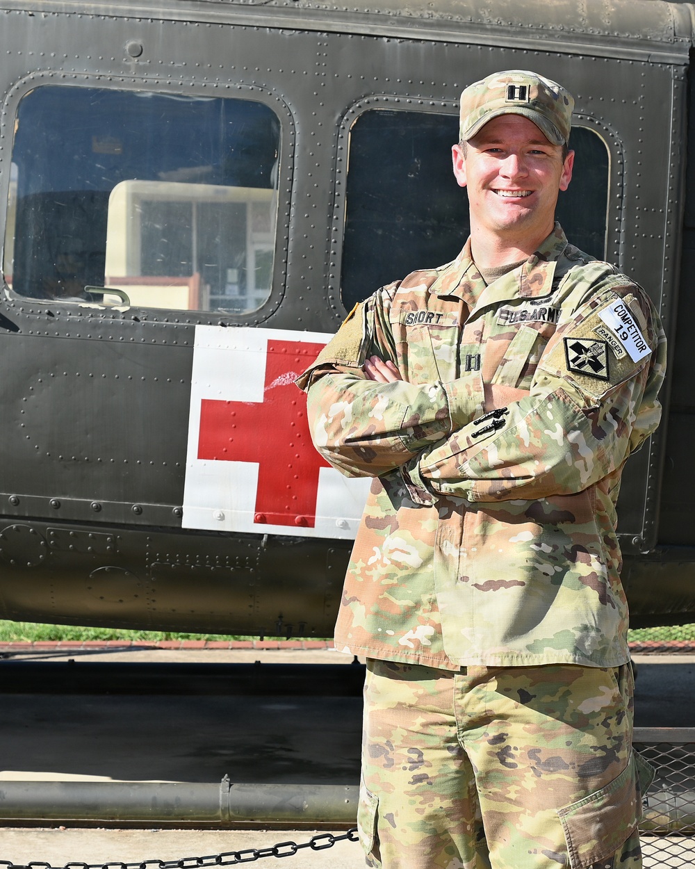 A U.S. Army Captain in OCP (Operational Camouflage Pattern) uniform stands with arms crossed in front of a military medical helicopter marked with a Red Cross symbol. The soldier is smiling and wearing multiple patches including a Ranger tab, U.S. Army tape, a unit patch on the right shoulder, and a 'Competitor 19' patch. The setting appears to be an outdoor display or static exhibit area.