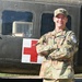 A U.S. Army Captain in OCP (Operational Camouflage Pattern) uniform stands with arms crossed in front of a military medical helicopter marked with a Red Cross symbol. The soldier is smiling and wearing multiple patches including a Ranger tab, U.S. Army tape, a unit patch on the right shoulder, and a 'Competitor 19' patch. The setting appears to be an outdoor display or static exhibit area.