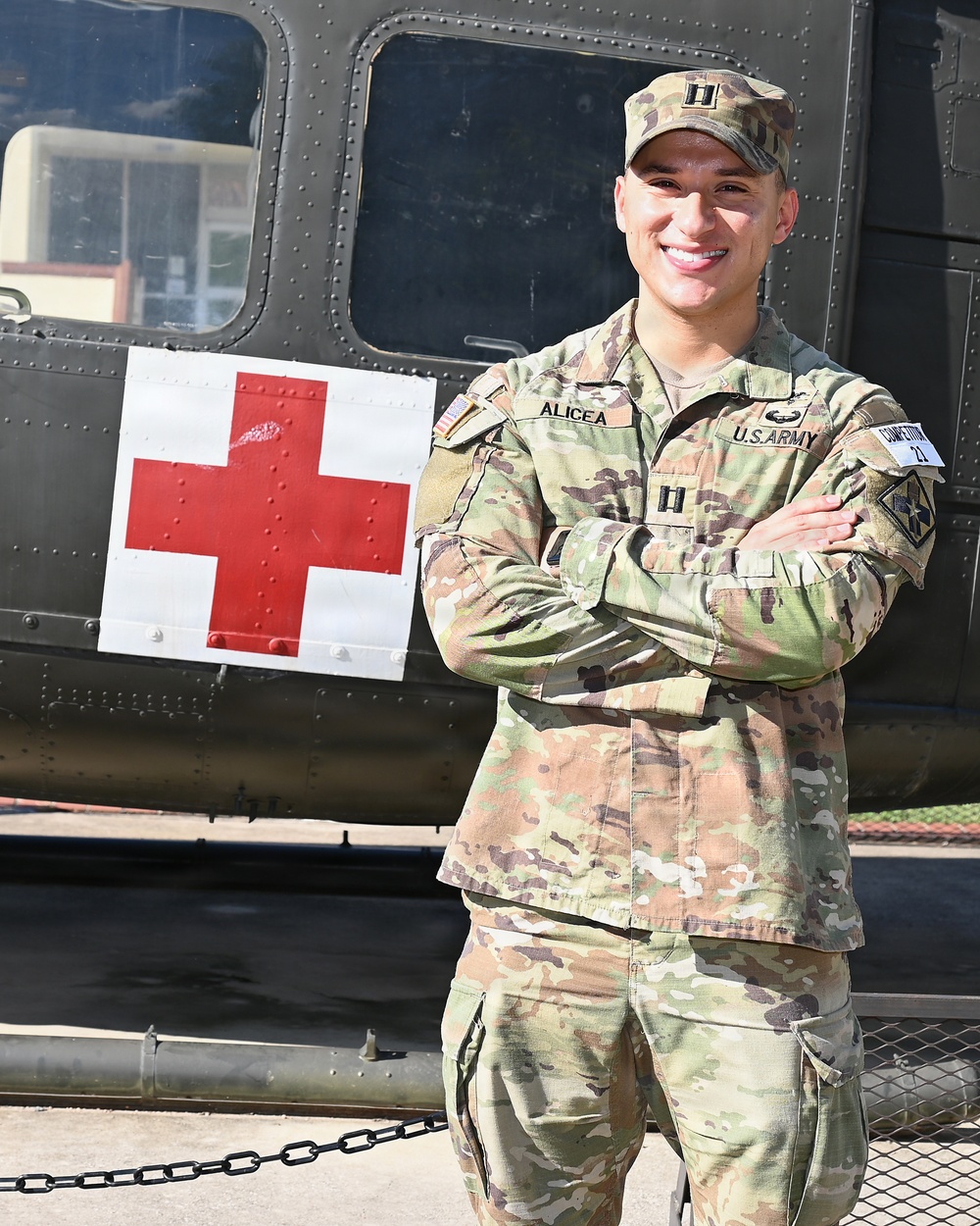 A U.S. Army Captain named Alicea stands confidently with arms crossed in front of a military medevac helicopter marked with a Red Cross symbol. He is wearing the Operational Camouflage Pattern (OCP) uniform with Captain rank insignia (two silver bars) visible on his chest. The setting appears to be an outdoor military display or installation.