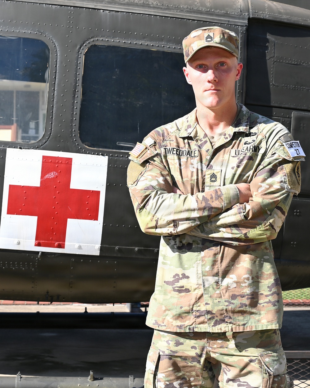 A U.S. Army Staff Sergeant named Tweeddale stands at attention with arms crossed in front of a military medevac helicopter marked with a Red Cross symbol. He is wearing Operational Camouflage Pattern (OCP) uniform with visible rank insignia, name tape, U.S. Army tape, and a competition number patch on his right sleeve. The image appears to be an official or semi-official military portrait photograph.