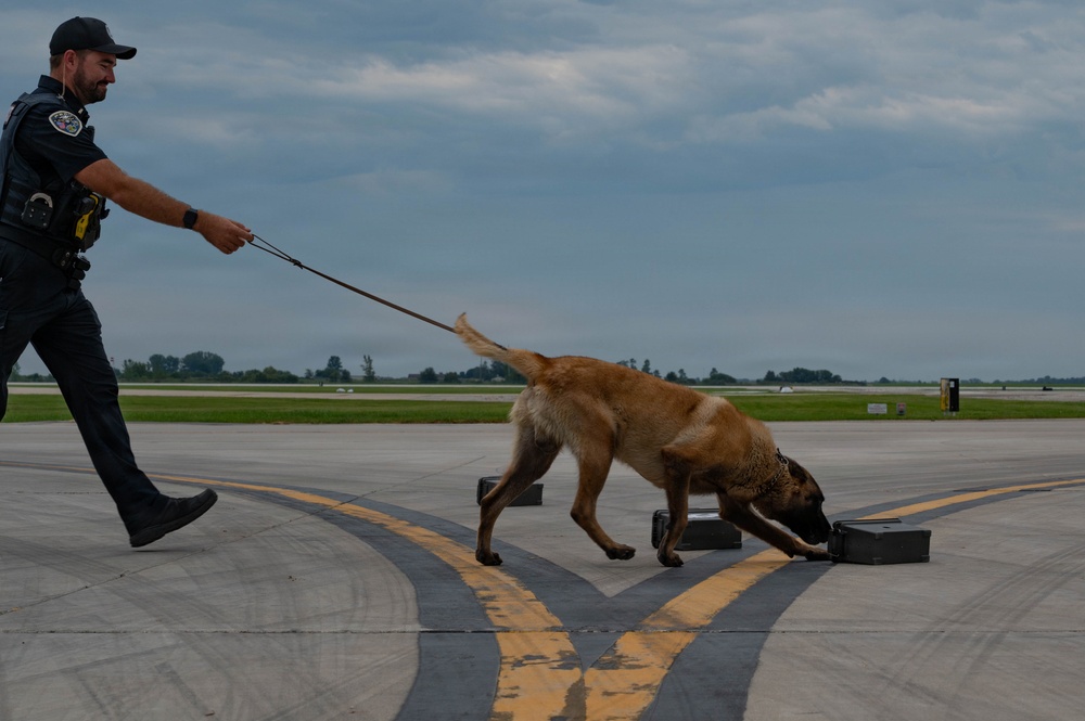 A law enforcement officer in dark tactical uniform is conducting a K9 detection exercise on an airport tarmac. A Belgian Malinois on a leash is actively sniffing a set of dark-colored cases or boxes placed on the runway surface. The setting appears to be a controlled training or demonstration scenario at an airfield.
