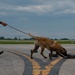 A law enforcement officer in dark tactical uniform is conducting a K9 detection exercise on an airport tarmac. A Belgian Malinois on a leash is actively sniffing a set of dark-colored cases or boxes placed on the runway surface. The setting appears to be a controlled training or demonstration scenario at an airfield.