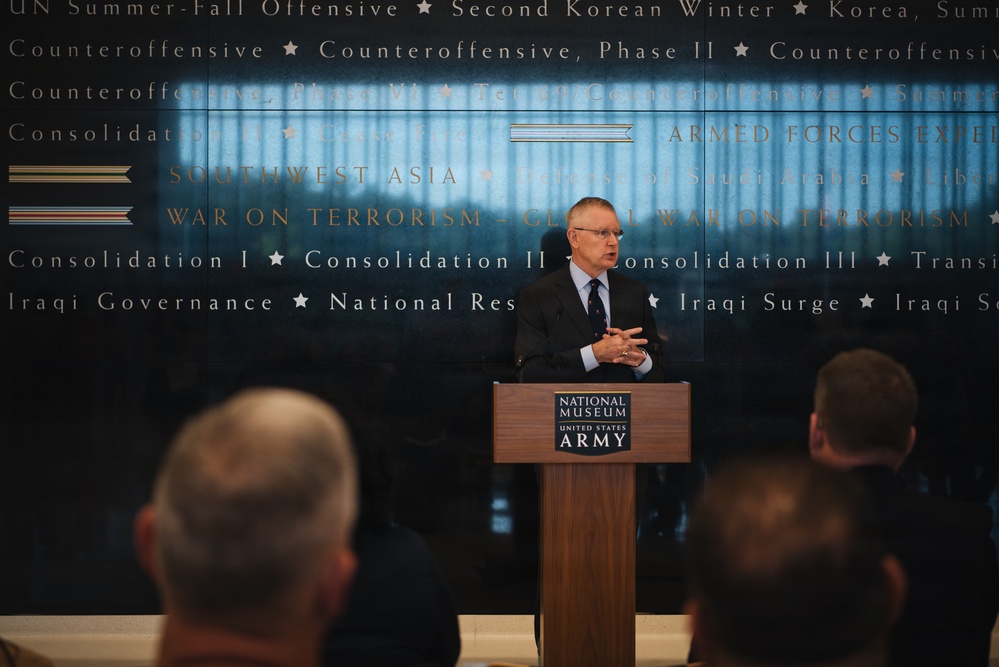 A man in a dark suit is speaking at a podium bearing the 'National Museum United States Army' logo, addressing an audience. The background features a large display wall with military campaign names and service ribbon graphics, including references to Southwest Asia, War on Terrorism, Korean War operations, and Iraqi campaigns. The setting appears to be a formal event or ceremony at the National Museum of the United States Army.