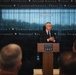 A man in a dark suit is speaking at a podium bearing the 'National Museum United States Army' logo, addressing an audience. The background features a large display wall with military campaign names and service ribbon graphics, including references to Southwest Asia, War on Terrorism, Korean War operations, and Iraqi campaigns. The setting appears to be a formal event or ceremony at the National Museum of the United States Army.