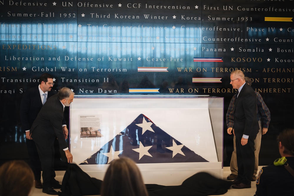 Three men are presenting a folded American flag displayed in a glass case at what appears to be a military museum or memorial. The flag is ceremonially folded in the traditional triangular shape, showing white stars on a blue field. The backdrop features a large wall inscribed with military campaign names and unit citations spanning multiple conflicts from Korea through the War on Terrorism.