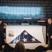 Three men are presenting a folded American flag displayed in a glass case at what appears to be a military museum or memorial. The flag is ceremonially folded in the traditional triangular shape, showing white stars on a blue field. The backdrop features a large wall inscribed with military campaign names and unit citations spanning multiple conflicts from Korea through the War on Terrorism.