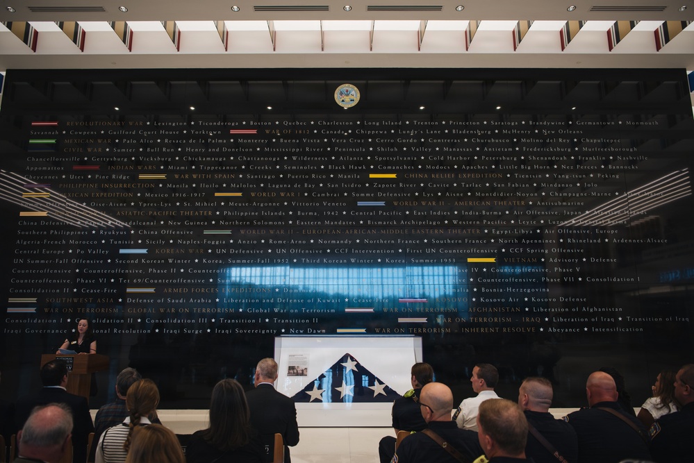 A formal ceremony is taking place in what appears to be a military museum or memorial hall, featuring a large dark wall inscribed with the names of U.S. Army campaigns and battles spanning from the Revolutionary War to modern conflicts. A speaker stands at a podium on the left while an audience is seated facing a folded American flag displayed on a table or stand at the center. The setting suggests a dedication, memorial, or official military ceremony.