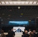 A formal ceremony is taking place in what appears to be a military museum or memorial hall, featuring a large dark wall inscribed with the names of U.S. Army campaigns and battles spanning from the Revolutionary War to modern conflicts. A speaker stands at a podium on the left while an audience is seated facing a folded American flag displayed on a table or stand at the center. The setting suggests a dedication, memorial, or official military ceremony.