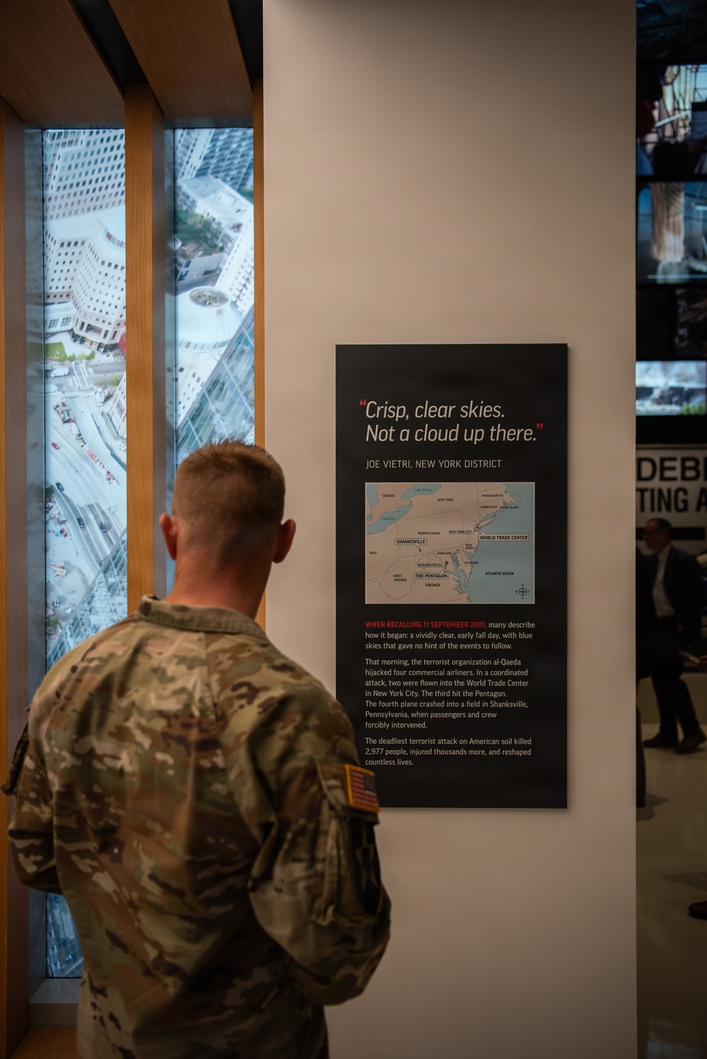 A military service member in camouflage uniform stands with their back to the camera, reading an exhibit panel at what appears to be the 9/11 Memorial Museum. The panel features a quote 'Crisp, clear skies. Not a cloud up there.' attributed to Joe Vietri, New York District, along with a map showing the attack locations and descriptive text about September 11, 2001. The setting includes floor-to-ceiling windows with an aerial view of lower Manhattan visible in the background.
