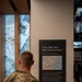 A military service member in camouflage uniform stands with their back to the camera, reading an exhibit panel at what appears to be the 9/11 Memorial Museum. The panel features a quote 'Crisp, clear skies. Not a cloud up there.' attributed to Joe Vietri, New York District, along with a map showing the attack locations and descriptive text about September 11, 2001. The setting includes floor-to-ceiling windows with an aerial view of lower Manhattan visible in the background.