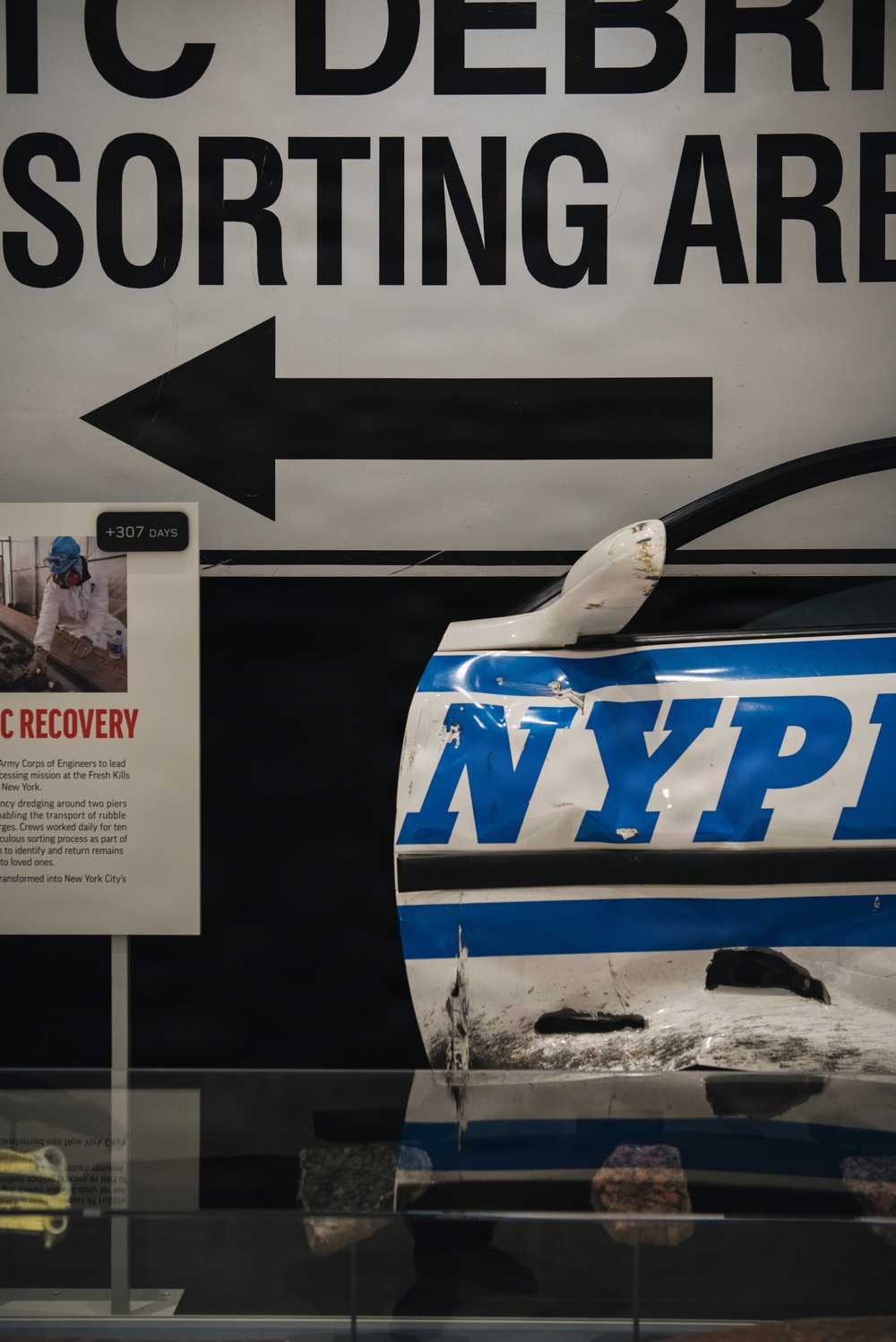 This image shows a museum exhibit, likely at the 9/11 Memorial Museum in New York City. The display features a heavily damaged NYPD police vehicle door/panel alongside a sign reading 'WTC DEBRIS SORTING AREA' with a directional arrow. An informational placard marked '+307 DAYS' references recovery operations at Fresh Kills landfill involving the Army Corps of Engineers.