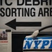 This image shows a museum exhibit, likely at the 9/11 Memorial Museum in New York City. The display features a heavily damaged NYPD police vehicle door/panel alongside a sign reading 'WTC DEBRIS SORTING AREA' with a directional arrow. An informational placard marked '+307 DAYS' references recovery operations at Fresh Kills landfill involving the Army Corps of Engineers.