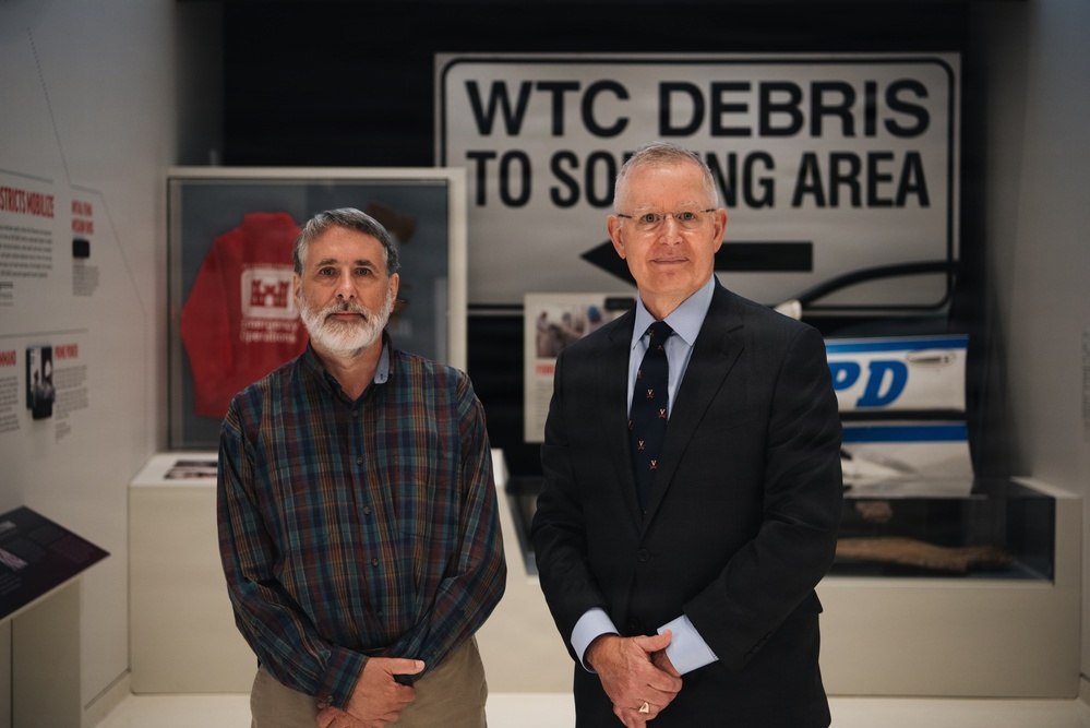 Two men are standing together in what appears to be a museum or memorial exhibition related to the September 11, 2001 World Trade Center attacks. Behind them is a large directional sign reading 'WTC DEBRIS TO SORTING AREA,' along with display cases containing artifacts including a red jacket and what appears to be an NYPD vehicle or panel. The setting suggests this is a commemorative or historical exhibit.