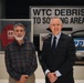 Two men are standing together in what appears to be a museum or memorial exhibition related to the September 11, 2001 World Trade Center attacks. Behind them is a large directional sign reading 'WTC DEBRIS TO SORTING AREA,' along with display cases containing artifacts including a red jacket and what appears to be an NYPD vehicle or panel. The setting suggests this is a commemorative or historical exhibit.