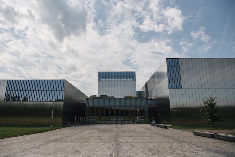 A modern architectural complex featuring multiple large rectangular structures clad in reflective glass and dark metallic panels. The buildings are arranged symmetrically around a central entrance, with a wide paved plaza leading to the main entry. The design is contemporary and minimalist, set against a partly cloudy sky.