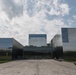 A modern architectural complex featuring multiple large rectangular structures clad in reflective glass and dark metallic panels. The buildings are arranged symmetrically around a central entrance, with a wide paved plaza leading to the main entry. The design is contemporary and minimalist, set against a partly cloudy sky.