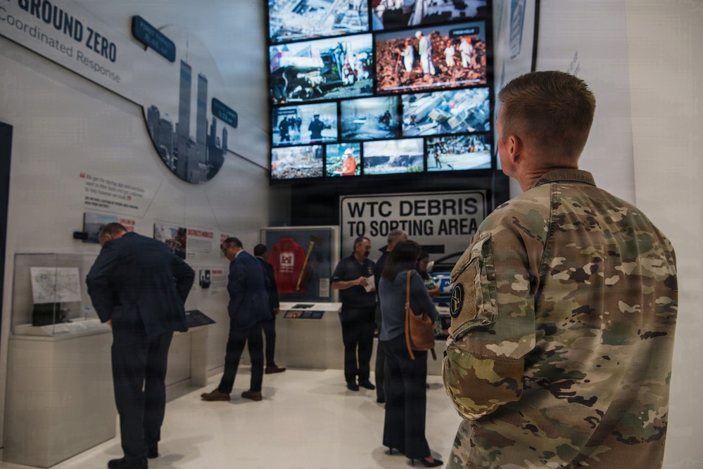 Visitors are touring what appears to be the 9/11 Memorial Museum, specifically a section dedicated to Ground Zero and the coordinated response efforts. A military service member in MultiCam/OCP camouflage uniform is visible in the foreground, observing the exhibit. The display includes a large video wall showing archival footage, a 'WTC Debris to Sorting Area' sign, and various artifacts and informational panels.