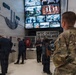 Visitors are touring what appears to be the 9/11 Memorial Museum, specifically a section dedicated to Ground Zero and the coordinated response efforts. A military service member in MultiCam/OCP camouflage uniform is visible in the foreground, observing the exhibit. The display includes a large video wall showing archival footage, a 'WTC Debris to Sorting Area' sign, and various artifacts and informational panels.