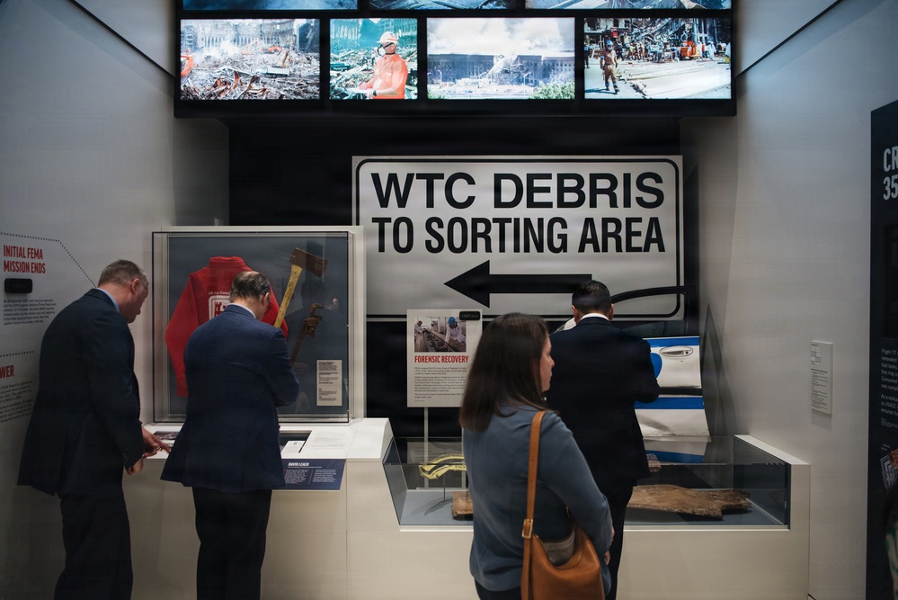 The image shows visitors examining exhibits at what appears to be the 9/11 Memorial Museum in New York City. A prominent directional sign reading 'WTC DEBRIS TO SORTING AREA' serves as a central artifact, surrounded by recovery-related items including a red jacket, tools, and debris fragments. Video screens above display archival footage of rescue and recovery operations.
