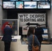 The image shows visitors examining exhibits at what appears to be the 9/11 Memorial Museum in New York City. A prominent directional sign reading 'WTC DEBRIS TO SORTING AREA' serves as a central artifact, surrounded by recovery-related items including a red jacket, tools, and debris fragments. Video screens above display archival footage of rescue and recovery operations.