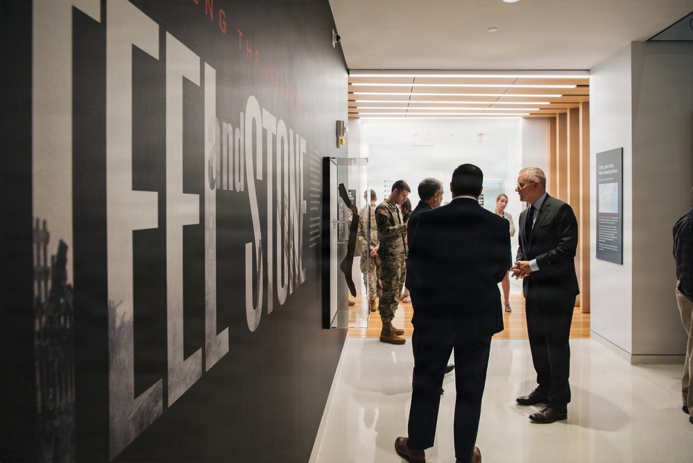 A group of people are gathered in a modern museum or exhibition corridor featuring large typographic wall graphics reading 'STEEL and STONE' (partial text visible). The visitors include individuals in business attire and at least one person in military camouflage uniform, suggesting an official or formal visit. The space features contemporary design with wood paneling, linear ceiling lights, and polished floors.