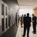 A group of people are gathered in a modern museum or exhibition corridor featuring large typographic wall graphics reading 'STEEL and STONE' (partial text visible). The visitors include individuals in business attire and at least one person in military camouflage uniform, suggesting an official or formal visit. The space features contemporary design with wood paneling, linear ceiling lights, and polished floors.