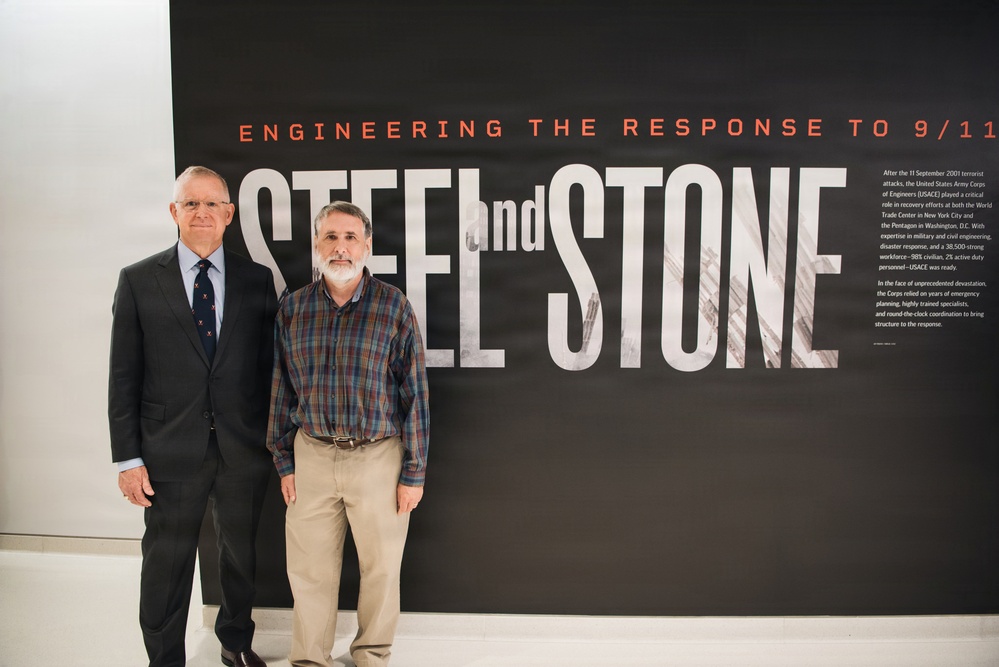 Two men are standing in front of a large exhibition display titled 'Engineering the Response to 9/11: Steel and Stone,' which appears to be a museum or commemorative exhibit about the U.S. Army Corps of Engineers' role in the 9/11 recovery efforts. One man is dressed in a formal dark suit with a tie, while the other wears a casual plaid shirt and khaki pants. The exhibit panel includes descriptive text about USACE's involvement at the World Trade Center and Pentagon sites.